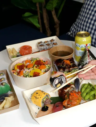 Table spread of sushi boxes, a chirashi bowl, edamame and a yuzu sparkling drink